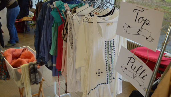 Kleiderstange mit nach Farben geordneten Kleidern darauf, ausgestattet mit schön geschriebenen Schildern "Top 2 Euro", "Pulli 4 Euro"