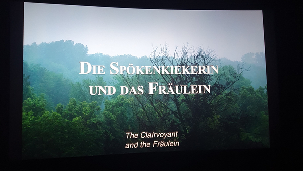 Auf der Kinoleinwand ist das Anfangsbild des Kurzfilms "Die Spökenkiekerin und das Fräulein" zu sehen. Der Titel steht in weißen Großbuchstaben mittig im Bild, darunter in klein der Titel in englisch. Im Hintergrund ist ein Wald und Hügel und Nebel zu sehen.
