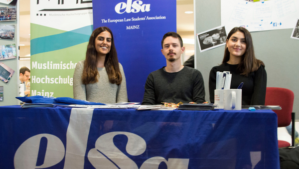 Drei Studierende sitzen vor einem Tisch. Er ist mit einem blauen Banner bedeckt, auf dem "elsa" steht, wobei das "s" als Paragraphenzeichen dargestellt wird.