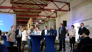 Das Bild zeigt zehn heitere Menschen vor Publikum, die sich im Rahmen einer Bierdeckelfragenrunde angeregt unterhalten.