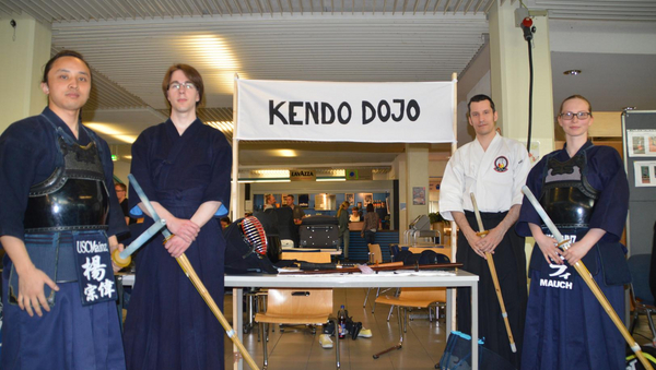 Vier Personen mit Kampfsport-Anzügen und Holzschwertern vor dem "Kendo Dojo"-Infostand