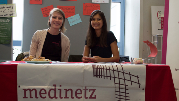 Zwei Studierende sitzen hinter einem Tisch, auf dem das Banner von "Medinetz Mainz" und einige Snacks zu sehen sind.