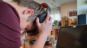 Ein junger Mann hält den Kopf vor einem Laptop gebeugt, während er sich das Headset vom Kopf nimmt.
