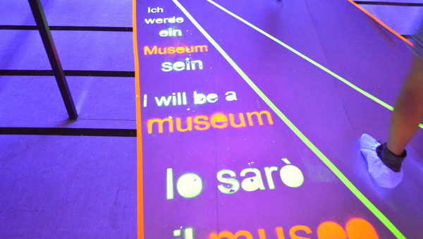 Der Boden ist in blaues Licht getaucht. Darin steht in grüner und roter leuchtender Schrift "Ich werde ein Museum sein" in verschiedenen Sprachen.