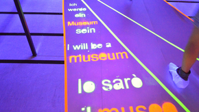 Der Boden ist in blaues Licht getaucht. Darin steht in grüner und roter leuchtender Schrift "Ich werde ein Museum sein" in verschiedenen Sprachen.
