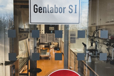 An einer Glastür prangt ein Schild mit der Aufschrift "Genlabor S1". Dahinter sind allerlei Gerätschaften zu sehen.