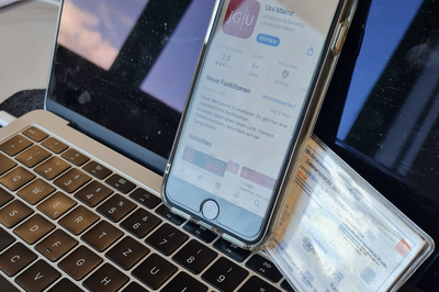 Ein Smartphone und ein Studierendenausweis lehnen an einem aufgeklappten Laptopbildschirm. Das Handy zeigt die JGU-App im App Store.