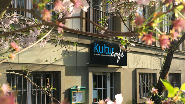 Im Vordergrund sieht man, unscharf, rosa Blüten. Im Hintergrund ein Gebäude und ein schwarzes Schild, auf dem mit blauer und weißer Schrift "Kulturcafé" steht.