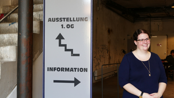 Annika Heinze steht rechts neben einem Schild, das mit einem Pfeil nach oben den Weg zu einer Ausstellung im ersten Obergeschoss zeigt. Ein weiterer Punkt auf dem Schild ist die "Information", die einem zweiten Pfeil nach rechts zu finden ist - da, wo Annika steht.