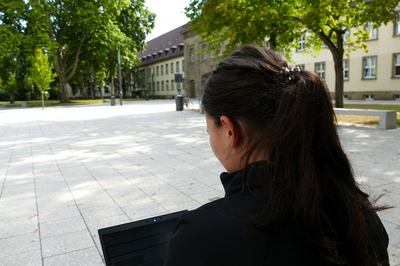 Eine Studentin sitzt vor dem Eingangsportal der JGU. Der Blick wandert über ihre Schulter zum Bildschirm des aufgeklappten Laptops.
