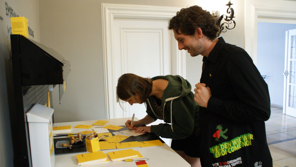 Eine Frau und ein Mann vor einem Tisch mit vielen gelben Zetteln. Die Frau schreibt auf einen Zettel.