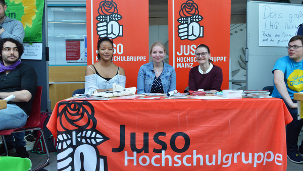 An einem Tisch sitzen drei junge Frauen. Am Tisch hängt vorn ein rotes Banner herunter, im Hintergrund stehen zwei rote Aufsteller.