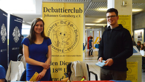 Zwei Personen stehen hinter einem Tisch mit roter Tischdecke. Beide halten Flyer in den Händen. Im Hintergrund steht ein großes gelbes Banner.