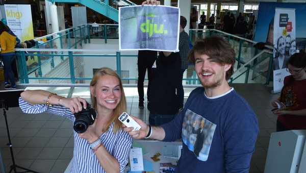Zwei Personen stehen im vorderen Bereich des Bildes. Sie halten eine Kamera und ein Tonaufnahmegerät in der Hand. Im Hintergrund hält eine Person ein Plakat hoch.