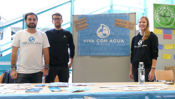 Drei Mitglieder der Organisation "Viva con Agua" präsentieren ihren Infostand, hinter dem eine Stellwand mit dem blauen Banner der Organisation platziert wurde.
