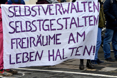 Ein weißer Banner auf dem in lilafarbener Schrift steht "Selbstverwalten, Selbstgestalten, Freiräume am Leben halten!"