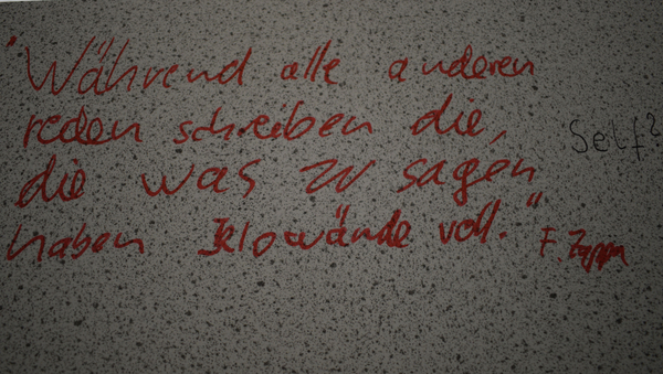 Eine weiße Fläche mit grauen Punkten. Darauf in roter Schrift ein Spruch über die Beschriftung von Klowänden.