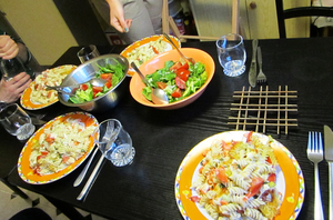 Ein dunkler Tisch, gedeckt für vier Personen mit Tellern, Besteck und Gläsern. Auf den Tellern ist Nudelsalat. In der Mitte stehen zwei Schalen mit grünem Salat. 