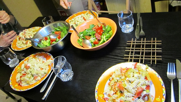 Ein dunkler Tisch, gedeckt für vier Personen mit Tellern, Besteck und Gläsern. Auf den Tellern ist Nudelsalat. In der Mitte stehen zwei Schalen mit grünem Salat. 