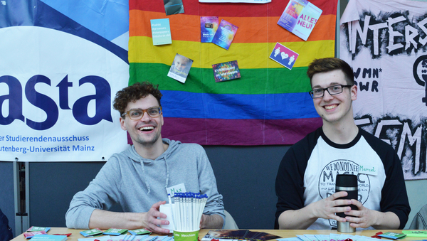 Zwei Männer sitzen hinter Tisch mit Infomaterial. Im Hintergrund hängt eine Regenbogenfahne.