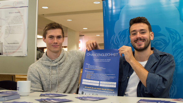 Zwei Studierende sitzen hinter einem Tisch mit Flyern und halten ein Plakat von "Chancengleichheit Mainz e.V." zwischen sich hoch.