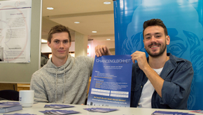 Zwei Studierende sitzen hinter einem Tisch mit Flyern und halten ein Plakat von "Chancengleichheit Mainz e.V." zwischen sich hoch.