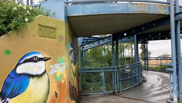 Neben dem gewundenen Aufgang einer Brücke ist links ein Graffiti zu sehen, das einen Vogel zeigt.