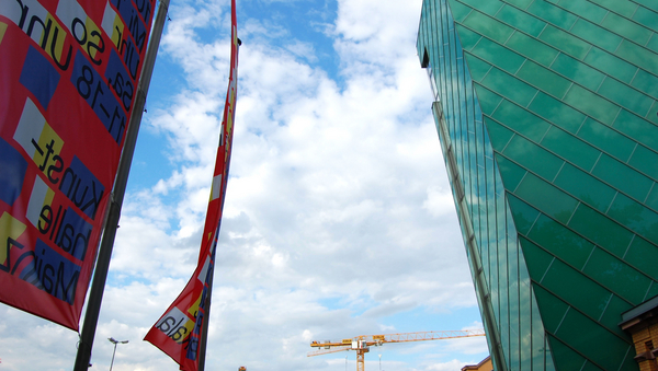 Rechts ein Gebäude, links rote Flaggen, die für eine Ausstellung werben. Im Hintergrund blauer Himmel.