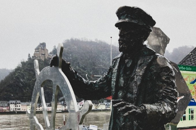 Die Statue eines Schifffahrerers mit Lenkrad ziert die Uferpromenade. Dahinter sieht man den Rhein und hohe Weinberge am gegenüberliegenden Ufer.