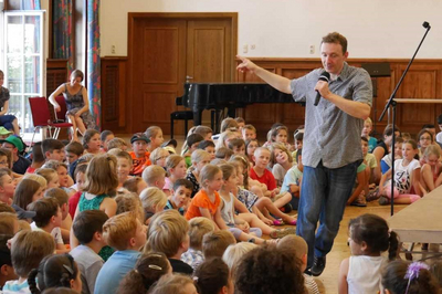Felix Koch spricht mit einem Mikrofon in der Hand zu einer großen Gruppe Kinder.