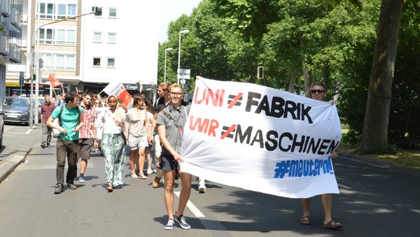 Demonstrationszug angeführt von zwei Männern, die ein Laken mit der Afschrift: "Uni≠Fabrik Wir≠Maschinen #meutern" tragen.