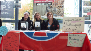 Drei Studierende sitzen hinter einem Tisch, an dem Schilder hängen, die ihre Arbeit und Angebote bewerben, etwa einen "antifaschistischen Stadtrundgang" oder andere Angebote zur politischen Bildung.