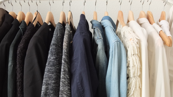 Kleiderstange, an der Kleidung hängt. Ganz links hängen dunkle Jacken und Pullis, dann graue, in der Mitte etwas Blaues, dann etwas Jeansfarbenes und ganz rechts hängt weiße Kleidung