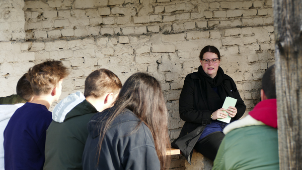 Annika Heinze sitzt mit mehreren Kindern aus einer Klasse vor einer Mauer auf dem Gelände der Gedenkstätte und erzählt ihnen etwas. Dabei helfen ihr auch die Karteikarten, die sie in ihren Händen hält.