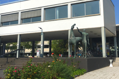 Das Philosophicum der Uni Mainz: Eine reduzierte Statue von einem Mann, der neben einem Pferd steht. Seid Blick geht gen blauer Himmel, danach streckt er auch die Hand aus. Es ist, als reckt er sich nach der feinen Wissenschaft. Davor befindet sich ein grünes, saftiges Blumenbeet. Es ist Sommer.