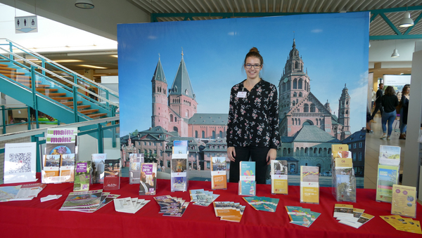Eine Vertreterin der Stadt Mainz steht vor einer Stellwand, die den Mainzer Dom zeigt. Vor ihr auf dem Tisch stehen und liegen zahlreiche Flyer mit Angeboten aus der Region Mainz.