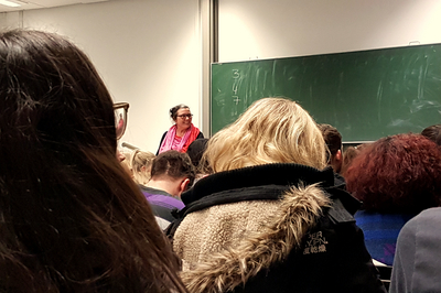 Prof. Dr. Sylvia Thiele steht in einem vollen Raum des Philosophicums vor einer Tafel, auf der die Zahlen 3, 4 und 7 geschrieben stehen.