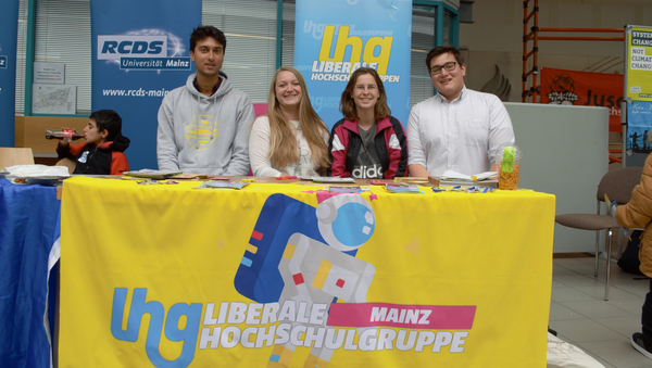 Die vier Studierenden der Liberalen Hochschulgruppe sitzen  hinter ihrem Tisch, der mit dem gelben Banner der politischen Gruppe versehen ist.