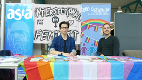 Zwei Vertreter des Schwulenreferats sitzen vor einem Tisch, der von verschiedenen bunten Flaggen bedeckt ist. Auf der Stellwand im Hintergrund hängen zwei Poster des Referats.