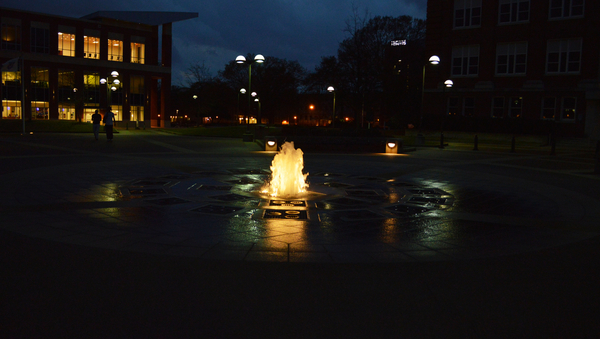 A picture taken by night. In the center a fountain which is in light. In the backround buildings and some lights. 