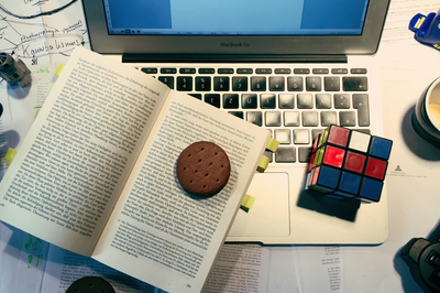Ein voller Schreibtisch: Texte sind verteilt, darauf steht ein Laptop. Darauf liegt ein Buch, auf dem ein Keks liegt. Daneben liegt ein Zauberwürfel, eine leere Kaffeetasse und ein Playstation Controller.