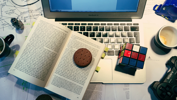 Ein voller Schreibtisch: Texte sind verteilt, darauf steht ein Laptop. Darauf liegt ein Buch, auf dem ein Keks liegt. Daneben liegt ein Zauberwürfel, eine leere Kaffeetasse und ein Playstation Controller.