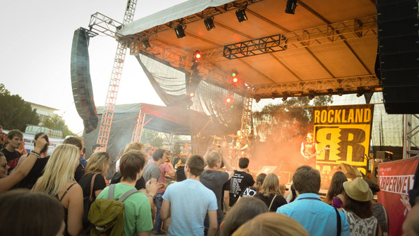 Man sieht eine Bühne, auf der eine Band spielt, inmitten von bunten Lichtern und Rauch. Hinten an der Bühne hängt ein großes, gelb-schwarzes Plakat, auf dem "Rockland" steht. Im Vordergrund sieht man die Zuschauer. 