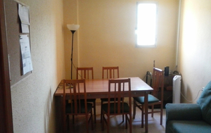 In der Mitte ein Tisch mit fünf Stühlen. Rechts oben ein Fenster. Rechts unten eine Couch. Links neben dem Tisch eine Wand.