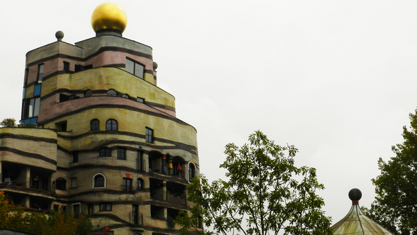 Links ein Gebäude mit Querstreifen und bunten Farben sowie einer goldenen Kugel auf der Spitze. Rechts ein Baum. 