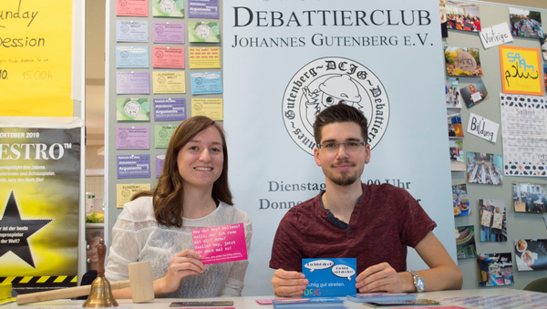 Zwei Studierende sitzen hinter einem Tisch, hinter dem sich das Banner des Debattierclubs befindet. Beide Student:innen halten Flyer hoch.