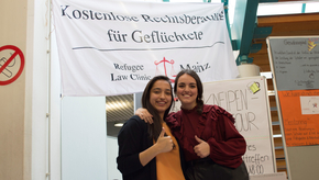Zwei Studierende stehen vor einem Banner der Kostenlosen Rechtsberatung für Geflüchtete, die alternativ auch als Refugee Law Clinic Mainz ausgewiesen wird.