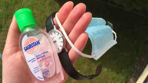 In einer geöffneten Hand liegen Desinfektionsmittel, eine Armbanduhr und eine OP-Maske.
