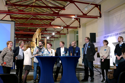 Das Bild zeigt zehn heitere Menschen vor Publikum, die sich im Rahmen einer Bierdeckelfragenrunde angeregt unterhalten.