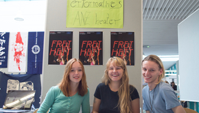 Drei Studierende sitzen vor einem Tisch, auf dem zahlreiche Flyer ausgelegt wurden. Auf der Stellwand hinter ihnen sind das Logo des Performativen Tanz-Theaters sowie die Poster ihrer Produktion "Freiheit" abgebildet.